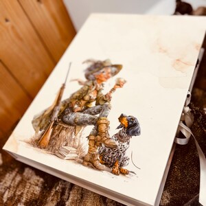 Könnte beinhalten: Eine rechteckige, cremefarbene Schachtel mit einer Aquarellillustration einer Person in Outdoor-Kleidung, die auf einem Baumstumpf sitzt, begleitet von einem Gewehr und einem gefleckten Hund. Die Schachtel hat einen weißen Bandverschluss.