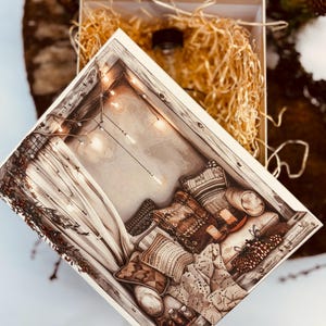 Könnte beinhalten: Eine quadratische Geschenkbox mit einem Deckel, der eine detaillierte Illustration einer gemütlichen Ecke mit Kissen, Decken und Lichterketten zeigt. Die offene Box enthält eine klare Glasflasche, eingebettet in helles, geschreddertes Füllmaterial.