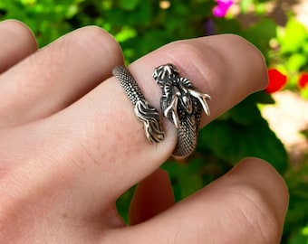 Op de afbeelding: Een zilveren drakenring met ingewikkelde details. De ring wikkelt zich om de vinger en heeft een drakenkop- en staartontwerp. De schubben en kenmerken van de draak zijn duidelijk gedefinieerd, waardoor een textuur ontstaat. De ring wordt op een vinger gedragen.