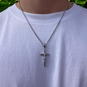 May include: A silver chain necklace with a silver cross pendant. The cross has a detailed design and a small figure on the front.