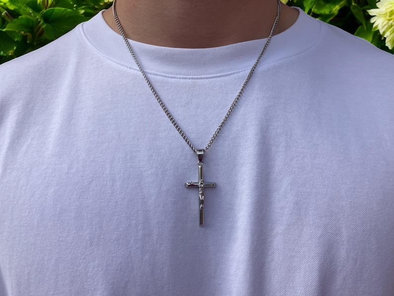May include: A silver chain necklace with a silver cross pendant. The cross has a detailed design and a small figure on the front.