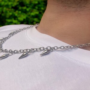 以下が含まれることがあります： 銀色のチェーンネックレスに、複数の銀色のスパイクがぶら下がっています。ネックレスは白いシャツを着た人が着用しています。