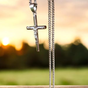 May include: A silver chain necklace with a silver cross pendant. The cross features a small figure of Jesus Christ. The necklace is hanging against a blurred background of a sunset and green trees.