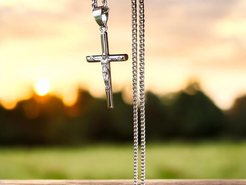 May include: A silver chain necklace with a silver cross pendant. The cross features a small figure of Jesus Christ. The necklace is hanging against a blurred background of a sunset and green trees.