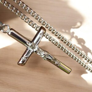 May include: A silver chain necklace with a silver cross pendant featuring a detailed depiction of a figure. The cross is attached to the chain with a ring.