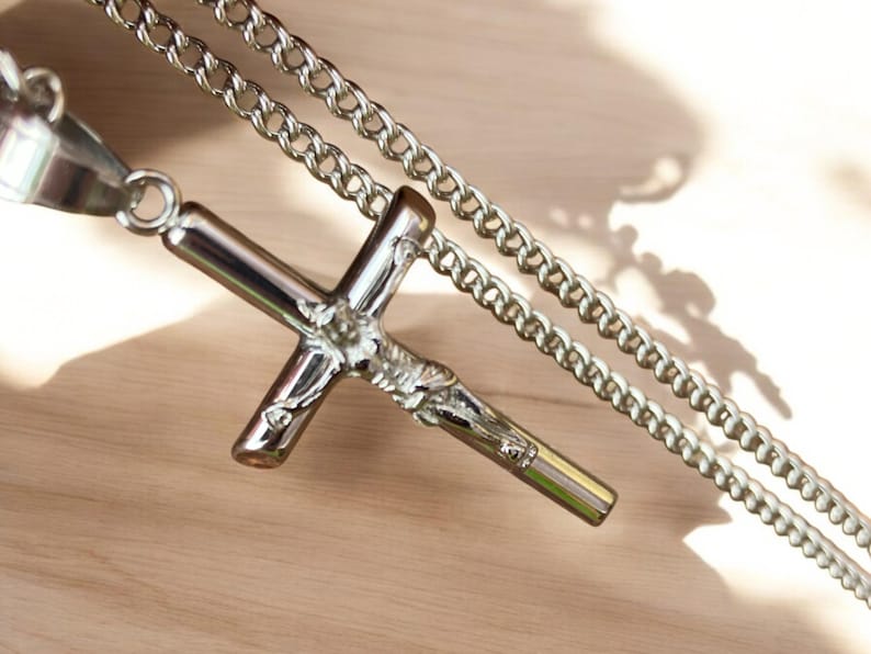 May include: A silver chain necklace with a silver cross pendant featuring a detailed depiction of a figure. The cross is attached to the chain with a ring.
