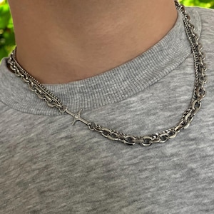 May include: A silver chain necklace with a small silver cross pendant. The necklace is layered with two different chain styles.