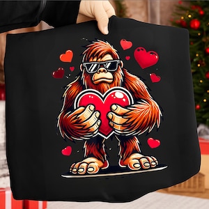 Op de afbeelding: Zwarte draagtas met een cartoon Bigfoot met een zonnebril en een rood hart. Rode harten zweven rond de Bigfoot. De Bigfoot is bruin en oranje met een zwarte omtrek. De tas wordt door een persoon vastgehouden.