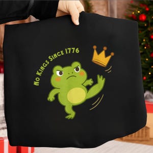 Op de afbeelding: Zwarte draagtas met een cartoonkikker die tegen een gouden kroon trapt. De kikker is groen met een gele buik en roze wangen. De tekst luidt "No Kings Since 1776" in een cirkel rond de kikker.