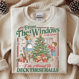 Op de afbeelding: Crèmekleurige sweatshirt met een vintage kerstafbeelding. Het ontwerp toont kinderen die een kerstboom versieren, een hond en de tekst "From The Windows To The Walls, I'm About To Deck These Halls."