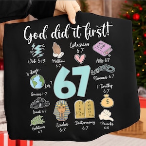 Op de afbeelding: Zwart t-shirt met de tekst "God did it first!" in witte letters. Het ontwerp bevat het nummer 67 in teal, samen met verschillende bijbelse verwijzingen en illustraties, zoals een hart, een wereldbol en een geldbuidel.