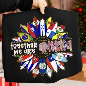 Puede incluir: Camiseta negra con un diseño de girasol compuesto por banderas de países americanos. El texto "together we are America" se muestra en blanco y letras coloridas. El diseño está centrado en la camiseta.