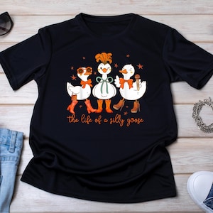 Op de afbeelding: Zwart T-shirt met een grillige illustratie van drie ganzen. De ganzen dragen accessoires zoals zonnebrillen, strikken en laarzen. De tekst "the life of a silly goose" staat eronder in het oranje.