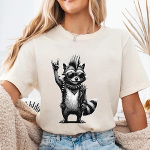 May include: Cream-colored t-shirt featuring a black and white illustration of a raccoon wearing sunglasses, a studded collar, and a leather jacket. The raccoon is making a rock and roll hand gesture.