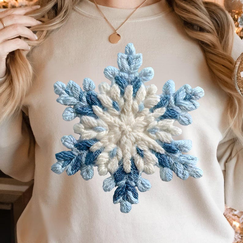 May include: A cream-coloured sweatshirt with a large, embroidered snowflake design. The snowflake is made with light blue, dark blue, and white threads, creating a textured effect. The design is centered on the chest of the garment.