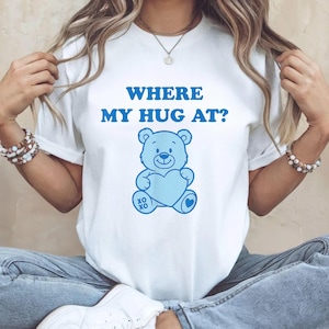 Pode incluir: Camiseta branca com o texto azul "WHERE MY HUG AT?". Um ursinho azul segurando um coração azul é impresso abaixo do texto. O urso tem as letras "XO XO" impressas abaixo.