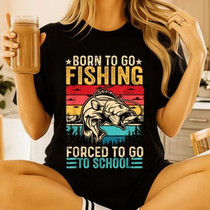 May include: Black t-shirt with a colorful graphic that reads "BORN TO GO FISHING FORCED TO GO TO SCHOOL." The graphic features a fish jumping out of the water, with a sunset and forest background. The shirt is short-sleeved.