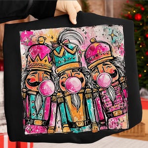 Op de afbeelding: Zwarte draagtas met een aquarel illustratie van drie notenkraker soldaten die roze kauwgom bellen blazen. De notenkrakers dragen kleurrijke uniformen en kronen, tegen een aquarel achtergrond met sterren.