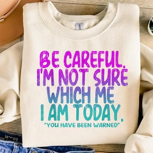 Könnte beinhalten: Cremefarbenes Sweatshirt mit dem Text "BE CAREFUL. I'M NOT SURE WHICH ME I AM TODAY. *YOU HAVE BEEN WARNED*" in Lila, Blau und Türkis. Das Sweatshirt ist gefaltet und auf einer hellen Oberfläche platziert.