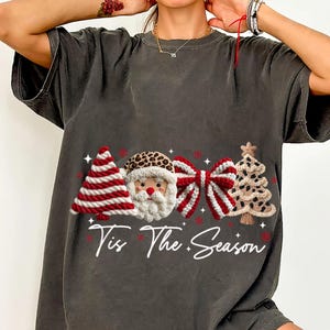 Op de afbeelding: Donkergrijs T-shirt met een kerstontwerp. Het heeft een rood-wit gestreepte kerstboom, een kerstman gezicht met een luipaardprint hoed, een rood-witte strik en een bruin gevlekte kerstboom. De tekst "Tis The Season" staat eronder.