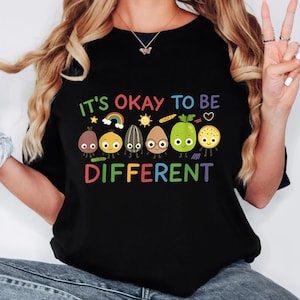 以下が含まれることがあります： 「IT'S OKAY TO BE DIFFERENT」という文字がカラフルにプリントされた黒いTシャツ。虹、太陽、様々な食べ物など、それぞれユニークなデザインの漫画のキャラクターが描かれています。