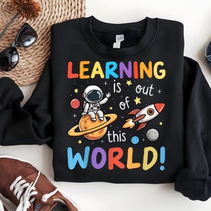 May include: Black sweatshirt with a colorful graphic that reads "LEARNING is out of this WORLD!" The design features an astronaut, planets, stars, and a rocket ship. The sweatshirt is a crew neck style.