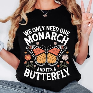 Op de afbeelding: Zwart T-shirt met een monarchvlinderontwerp. De tekst luidt: "WE ONLY NEED ONE MONARCH AND IT'S A BUTTERFLY". De vlinder is oranje en zwart, met bloemmotieven. Een casual top met een natuurthema.