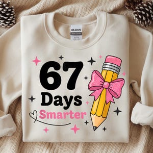 Op de afbeelding: Crèmekleurige sweatshirt met de tekst "67 Days Smarter" in zwart en roze. Een gele potlood met een roze gum en strik staat rechts. Roze sterren en zwarte sterren omringen de tekst en het potlood.