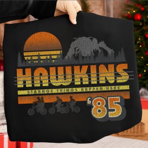 Op de afbeelding: Zwart T-shirt met een retro design. Het ontwerp toont het woord "Hawkins" in oranje en geel, een zonsondergang, een monstersilhouet en de tekst "Strange Things Happen Here" en "'85". Silhouetten van mensen op fietsen zijn ook inbegrepen.