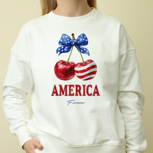 Op de afbeelding: Witte sweatshirt met een afbeelding van twee kersen, één met rood-witte strepen en de andere effen rood, vastgebonden met een blauwe strik met witte sterren. Het woord "AMERICA" is rood gedrukt, met "Forever" eronder.