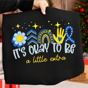 May include: Black tote bag with a colorful design featuring a blue and yellow flower, a rainbow, a hand, and a ribbon. The text "It's okay to be a little extra" is written in white and yellow.