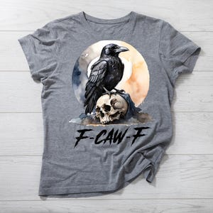 Op de afbeelding: Een grijs t-shirt met een aquarelontwerp van een zwarte raaf die op een schedel zit, tegen een maanverlichte achtergrond. De tekst "F-CAW-F" staat eronder gedrukt. Het t-shirt is gemaakt van een zacht materiaal.