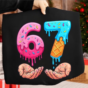 May include: Black t-shirt with a vibrant graphic of the numbers 67, styled as donuts and ice cream. The number 6 is pink with blue sprinkles, and the number 7 is blue with a waffle cone base. Two hands are shown below, also covered in sprinkles.