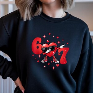 May include: Black sweatshirt featuring a red heart character wearing sunglasses and sneakers, dabbing between the numbers 6 and 7. Small red hearts and stars are scattered around the graphic. A fun, Valentine's Day themed design.