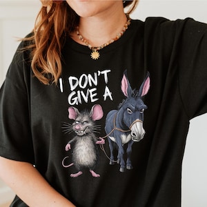 Peut inclure: T-shirt noir avec l'inscription "I DON'T GIVE A" au-dessus d'une illustration de dessin anim&eacute;. L'illustration repr&eacute;sente une souris grise aux oreilles roses tenant une corde attach&eacute;e &agrave; un &acirc;ne bleu fonc&eacute; &agrave; l'expression s&eacute;v&egrave;re.