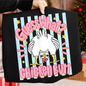 Op de afbeelding: Zwart T-shirt met een cartoon kip ontwerp. De tekst "Guess What? Chicken Butt" staat in roze bubbelletters. Het ontwerp bevat blauwe strepen, gele sterren en roze streepjes.