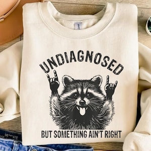 May include: Cream-colored sweatshirt with the text "UNDIAGNOSED" above a black and white raccoon graphic making a rock and roll hand gesture. Below the raccoon is the text "BUT SOMETHING AIN'T RIGHT".