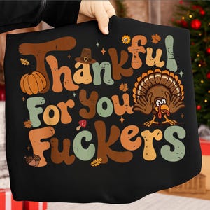 Può includere: T-shirt nera con grafica a tema Ringraziamento. Il design presenta le parole "Thankful For You Fuckers" in vari colori, insieme a una zucca, un tacchino e altre illustrazioni a tema autunnale.