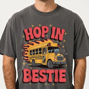 Op de afbeelding: Donkergrijze t-shirt met een gele schoolbus, vlammen en de tekst "HOP IN BESTIE". De bus heeft de tekst "STRUGGLE BUS" op de zijkant. Het ontwerp bevat sterren.