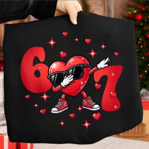 May include: Black graphic tee featuring a red heart character wearing sunglasses and sneakers, dabbing. The number 6 and 7 are in red, along with red hearts and stars. The design is on a black background.
