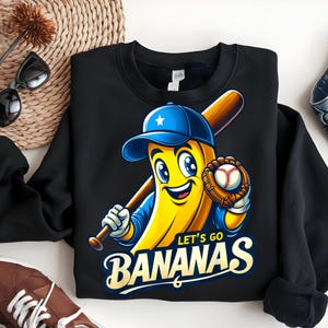 Op de afbeelding: Zwart sweatshirt met een cartoon banaan met een baseballpet en een knuppel en handschoen. De banaan heeft een lachend gezicht en de tekst "LET'S GO BANANAS" in gouden letters.