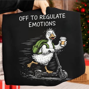 Puede incluir: Camiseta negra con un diseño de un pato de dibujos animados montando un scooter, con una mochila y un café. El texto "OFF TO REGULATE EMOTIONS" está impreso encima del pato.