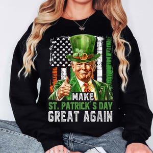 Op de afbeelding: Zwart sweatshirt met een afbeelding van een man die een groene leprechaun-hoed draagt en zijn duim omhoog steekt. De afbeelding is voor een Amerikaanse vlag met klavertjesvier. De tekst "Make St. Patrick's Day Great Again" is onder de afbeelding gedrukt.