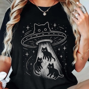 Op de afbeelding: Zwart T-shirt met een witte illustratie van een UFO die drie zwarte katten ontvoert. Het ontwerp bevat sterren en een kat die uit de UFO kijkt.