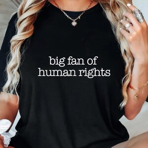 Puede incluir: Camiseta negra con el texto blanco "big fan of human rights". La camiseta es de color negro liso y tiene cuello redondo. El texto está en una fuente sencilla sin serifa. La camiseta es de estilo casual.