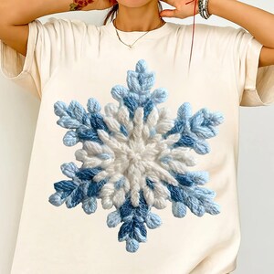 May include: A cream-coloured t-shirt with a large, textured snowflake design. The snowflake is made with white and blue yarn, creating a three-dimensional effect. The design is centred on the shirt, offering a winter theme.