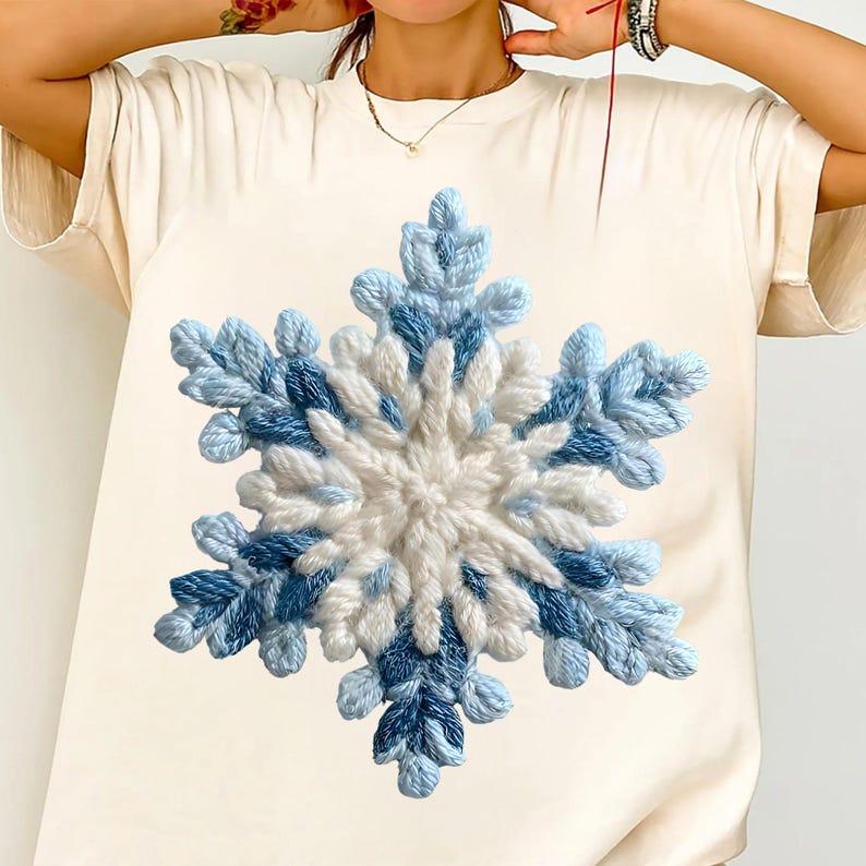 May include: A cream-coloured t-shirt with a large, textured snowflake design. The snowflake is made with white and blue yarn, creating a three-dimensional effect. The design is centred on the shirt, offering a winter theme.