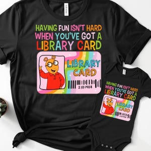 Op de afbeelding: Zwart t-shirt en babybody set met de tekst "HAVING FUN ISN'T HARD WHEN YOU'VE GOT A LIBRARY CARD". Beide items tonen een roze bibliotheekkaartafbeelding met een cartoonfiguur en barcode. De body is voor een baby.