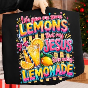 Op de afbeelding: Een zwart t-shirt met een kleurrijke afbeelding en de tekst Life gave me some LEMONS But my JESUS He be making LEMONADE. Het ontwerp bevat een glas gele limonade met een blauw-wit gestreept rietje, een roze strik en citroenen.