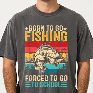 May include: Dark gray t-shirt with a colorful graphic. The design features a fish jumping out of the water, with the text "BORN TO GO FISHING" above and "FORCED TO GO TO SCHOOL" below. The graphic has a vintage, distressed look.